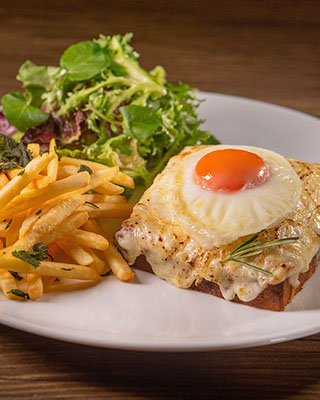 Croque Madame - Fatia de brioche, queijo emmental, presunto e molho bechamel servidos com batatas fritas, folhas orgânicas e ovo frito
